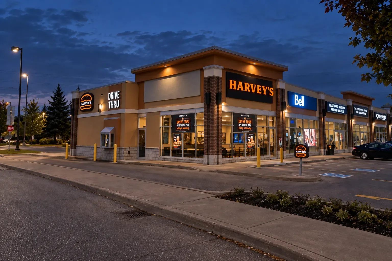 Harvey's restaurant with drive-thru at the end of a retail plaza, with EIFS and stucco storefronts lit at dusk